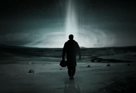 A lone figure stands under a beam of light on a desolate landscape at night
