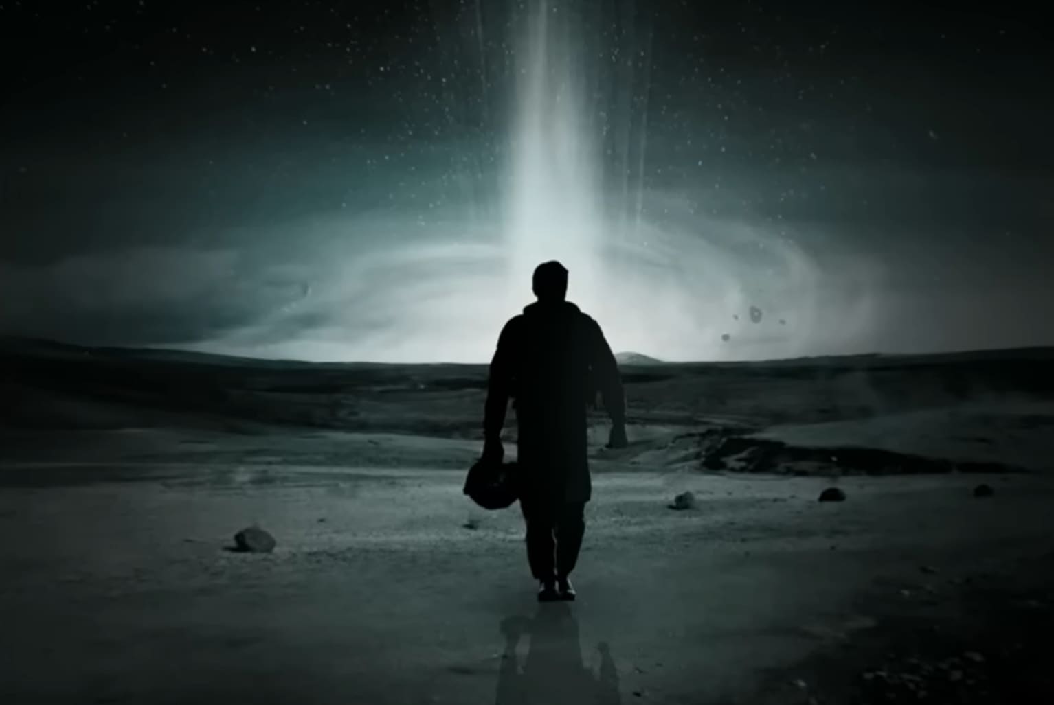 A lone figure stands under a beam of light on a desolate landscape at night